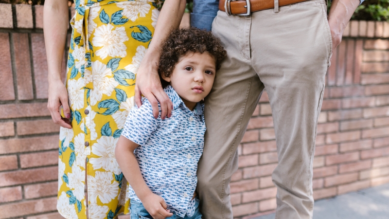 A Young Boy Stands Between His Parents, Holding onto His Mother's Leg While Looking Ahead