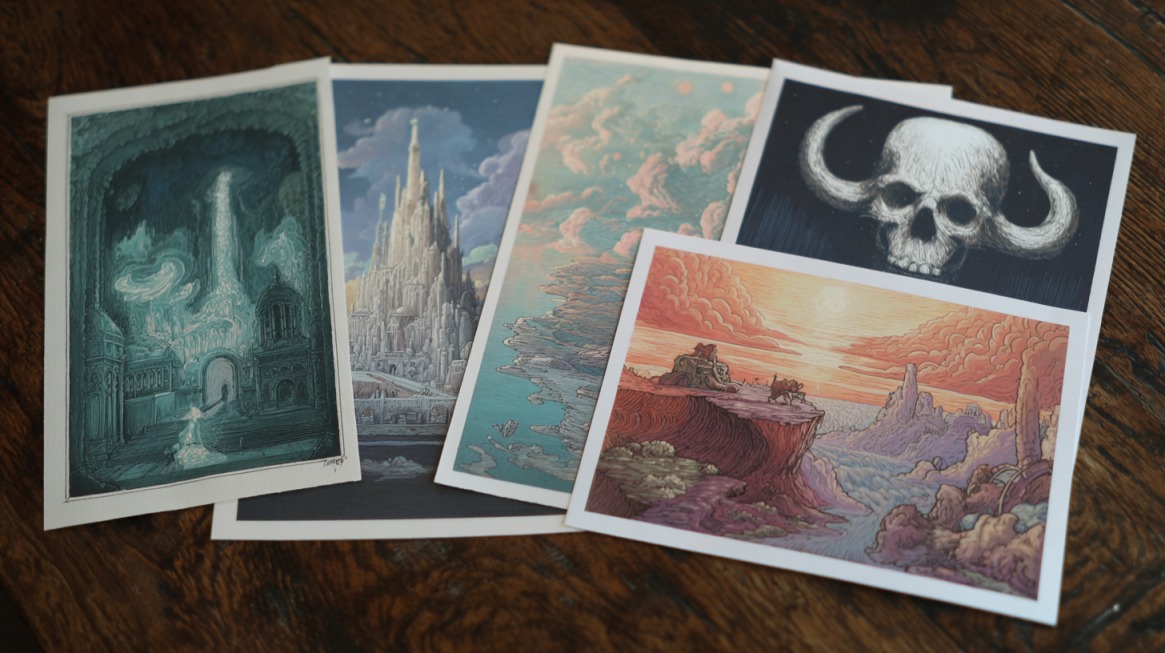 Five colorful fantasy-style art prints laid out on a wooden surface, featuring castles, landscapes, clouds, and a skull illustration