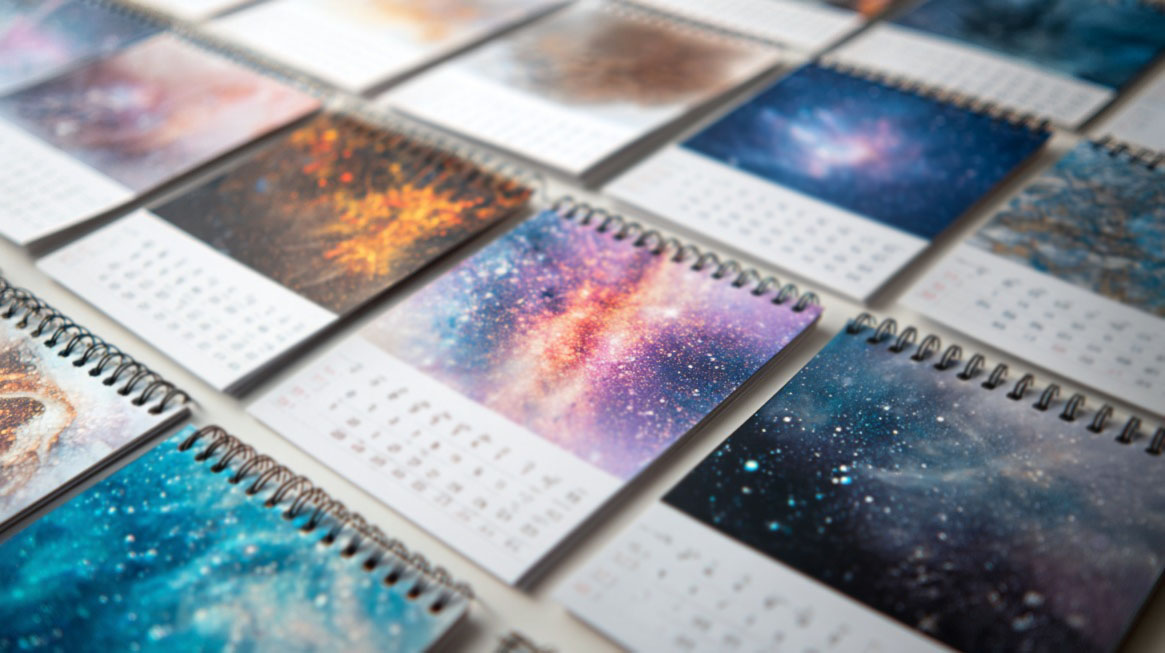 Multiple spiral-bound desk calendars laid out, each featuring vibrant space and galaxy-themed artwork above the monthly dates